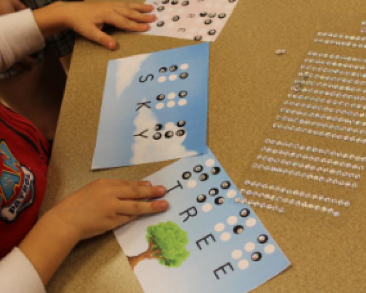 Child using sticky rhinestones to spell words in braille on illustrated cards